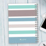 SparkGreetings® Array of Stripes Writing Journal, pastel-colored striped notebook with a spiral binding on a light wooden desk.