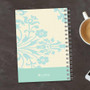 SparkGreetings® Tapestry Pattern Writing Journal, spiral-bound notebook with a light blue floral design on a cream background, placed on a dark wooden surface with a cup of coffee and a smartphone nearby.