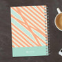 SparkGreetings® Tapestry Pattern Writing Journal, spiral-bound notebook with a vibrant, colorful tapestry-inspired cover design on a dark wooden surface next to a cup of coffee and a smartphone.