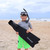 Child wearing Los Ninos Freedive Fins with snorkel gear, holding black fin bag on beach with ocean waves in background