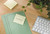 Post-it Recycled Z-Notes 76x76mm 100 Sheets Per Pad PEFC Canary Yellow (Pack 16), yellow sticky notes on a wooden desk with a plant and a keyboard.