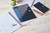 CW Office Oxford Notes Reporter Notebook, with blue and black covers, open and closed notebooks with lined pages, a silver paperclip, a black pencil, an orange pen, and a green mug on a light wooden desk.