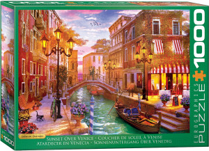 The artwork displays a narrow canal in Venice crisscrossed by a small arched stone bridge. The vibrant colors of a sunset cast a warm glow over the scene, with shades of orange, pink, and purple in the sky. Elegant street lamps are lit, casting light onto the canal and the buildings on either side. On the left, there is a row of historic buildings with visible balconies and ornate window frames. The building facades are painted in rich tones of red, orange, and brown, with ivy clinging to the walls. A small gondola is moored in the canal. On the right, a café with striped awnings and outdoor seating is visible. A couple stands on the bridge, admiring the view, while a cat prowls the walkway. The setting is lively yet serene, capturing the classic charm of Venice. The title, "Sunset over Venice" is printed along the bottom edge in white on green background. On the right hand edge, the company logo reads "EUROGRAPHICS PUZZLES" and the number ‘1000’ after it indicates the puzzle piece count.