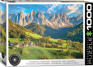 The image is a puzzle box that features a scenic landscape of the Italian Alps in the Dolomites. The foreground features a lush, green valley with scattered small buildings, including a church with a tall steeple surrounded by green fields and dense coniferous forests. In the background, a dramatic range of rocky, jagged peaks rises under a partly cloudy sky, illuminated by warm sunlight. The title, "Dolomites - Italian Alps" is printed along the bottom edge. On the right hand edge, the company logo reads "EUROGRAPHICS PUZZLES" in white text on black background and the number ‘1000’ after it indicates the puzzle piece count. In the bottom left, there is a picture of a camera lens with the words "HDR Photography - Scenic Landscape" printed on the inside rim of the lens.