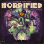 A board game titled "Horrified: World of Monsters," the first half of the title in large, all capital golden font across the top of the box and the second half much smaller beneath.  Below the title is a movie-poster-style arrangement of characters and elements atop a dark purple background.  The scene is, from bottom to top: A snarling creature with a human face, lion's claws, wings, and Pharoah's head and chin garb; a train with a fierce face on the front exiting a stone tunnel; a purple, three eyed creature with ram's horns and an ape-like face mid roar; a pale humanoid with white eyes, a long black braid, golden cap, and a slip of paper with chinese characters on it across the person's face; a tower with a large, bronze cog atop it; a being with an squid-like face, 5 amber eyes with slitted pupils, rough skin, and purple bat wings.