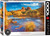 The image is a puzzle box with a striking photograph of Red Rock Crossing in Arizona, USA. The vivid landscape shows a winding river bordered by autumn foliage, with tall golden grasses and shrubs. In the background, iconic red rock formations rise against a vibrant blue sky dotted with scattered clouds. The foreground highlights the flowing water, reflecting the colorful scenery. The title, "Red Rock Crossing, Arizona" is printed along the bottom side. On the right hand edge, the company logo reads "EUROGRAPHICS PUZZLES" in white text on a black background and the number ‘1000’ after it indicates the puzzle piece count. In the bottom left, there is a picture of a camera lens with the words "HDR Photography - Scenic Landscape" printed on the inside rim of the lens.