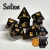A set of seven dark purple, sparkly polyhedral dice with golden numerals and cats on one side instead of numbers.  In the bottom right corner is a black cartoon cat wearing a pointy, wide-brimmed hat and sitting next to an orange, carved pumpkin.