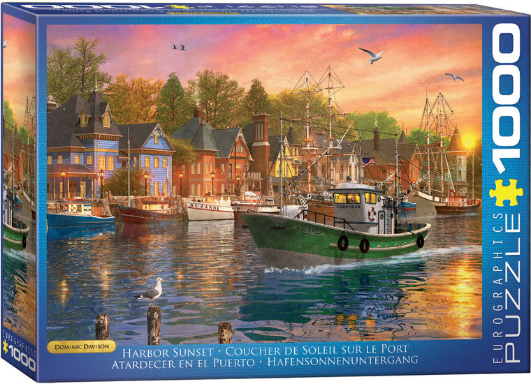 The image shows the box of a puzzle featuring the piece “Harbor Sunset” by Dominic Davison. The artwork depicts a vibrant harbor scene at sunset. Several fishing boats are anchored at the dock, with the prominent green boat in the foreground named "Cormoran." The water reflects the warm colors of the sunset sky, a blend of pink, orange, and purple hues. On the left, a quaint village with Victorian-style houses, painted in shades of blue and brick red, lines the waterfront. Trees with lush green foliage stand behind the buildings. A few birds, including a seagull, fly across the sky, while one seagull is perched on a wooden post in the foreground. Along the right hand edge  the company logo reads "EUROGRAPHICS PUZZLES" in white text on a blue background. The number ‘1000’ printed after it indicates the puzzle piece count. Along the bottom edge the title is printed in several different languages.