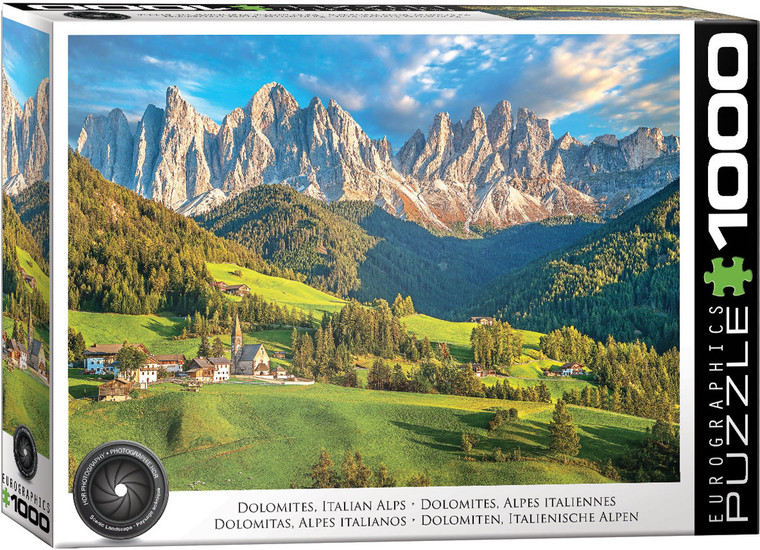 The image is a puzzle box that features a scenic landscape of the Italian Alps in the Dolomites. The foreground features a lush, green valley with scattered small buildings, including a church with a tall steeple surrounded by green fields and dense coniferous forests. In the background, a dramatic range of rocky, jagged peaks rises under a partly cloudy sky, illuminated by warm sunlight. The title, "Dolomites - Italian Alps" is printed along the bottom edge. On the right hand edge, the company logo reads "EUROGRAPHICS PUZZLES" in white text on black background and the number ‘1000’ after it indicates the puzzle piece count. In the bottom left, there is a picture of a camera lens with the words "HDR Photography - Scenic Landscape" printed on the inside rim of the lens.