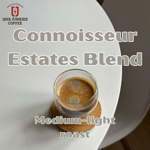 Java Junkies Connoisseur Estates Coffee. Clear class of coffee with cream sitting on a round coaster on a white table with a gray background.