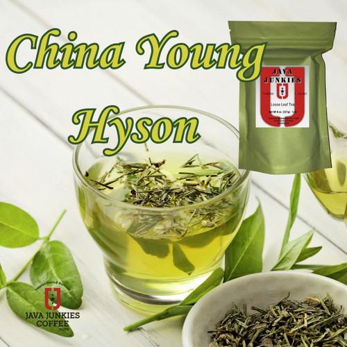 Clear glass cup of light green tea with loose leaves floating inside, next to a small white bowl of dried green tea on a white wooden surface.