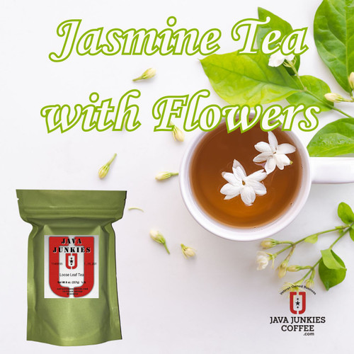 A white ceramic mug filled with amber tea garnished with fresh white jasmine flowers, surrounded by scattered green leaves and buds on a clean white surface.