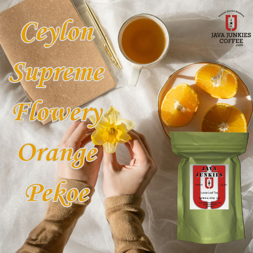 Hands holding a yellow flower near a notebook, a cup of tea, sliced oranges on a plate, and a green product bag on white fabric.