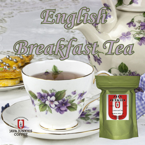 A floral teacup filled with amber tea rests on a matching saucer next to a decorative teapot, a plate of decorated cookies, and a green product package on a white tablecloth.
