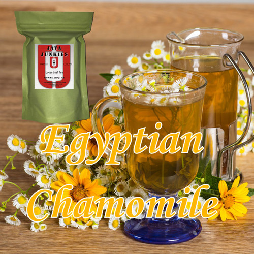 A decorative glass mug filled with golden chamomile tea and fresh daisy flowers resting next to a glass pitcher and scattered yellow wildflowers on a wooden table.