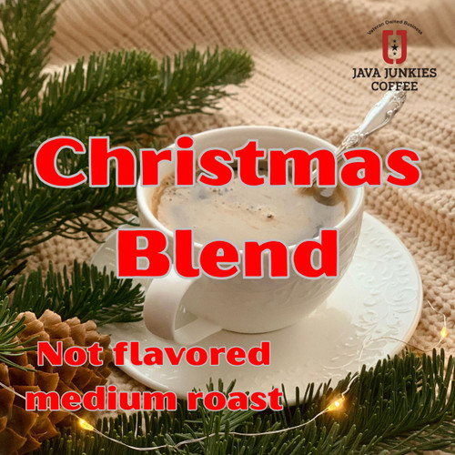 Java Junkies Christmas Coffee Blend. White coffee cup full of creamy coffee on a saucer with a spoon in it sitting next to a Christmas tree.