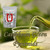 A transparent glass cup filled with bright green tea receiving a pour from a glass teapot, alongside a packaged pouch on a wooden table.