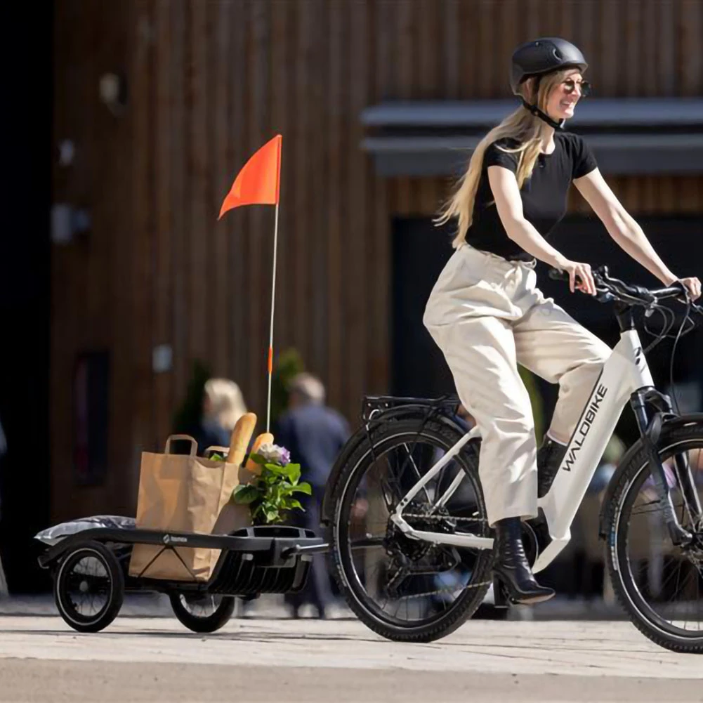 Woman cycling on a white e-bike with a Hamax bike trailer attached, featuring an orange safety flag and shopping bag, in an urban setting