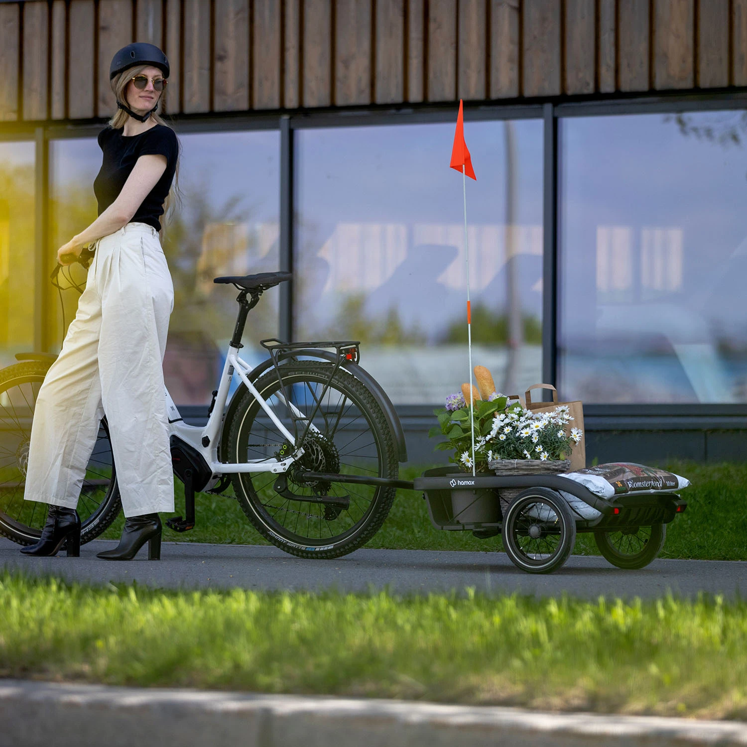 Woman with helmet and sunglasses standing next to a white e-bike with a Hamax bike trailer featuring an orange safety flag, on a sunny day with green grass and modern building