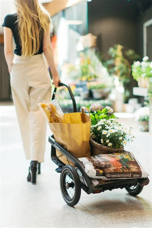 Hamax bike trailer loaded with flowers, a yellow tote bag, and Blomsterjord potting soil, being pulled through a flower market