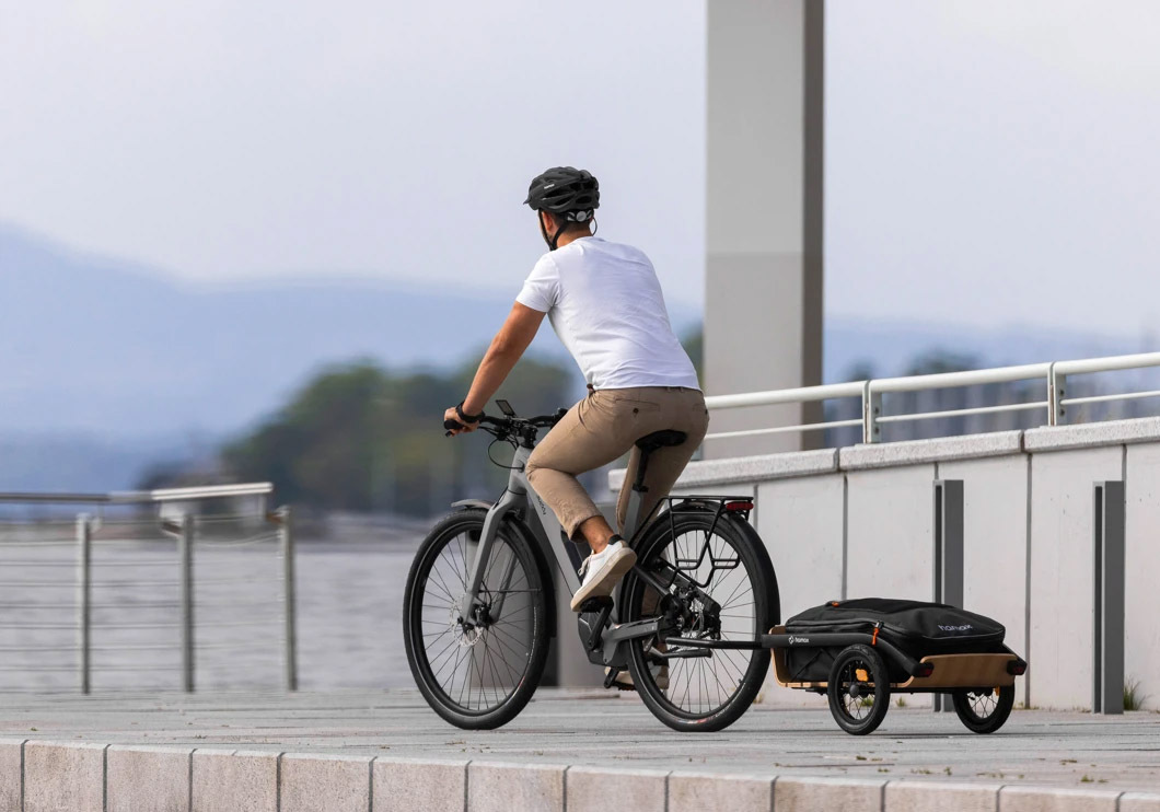 Person riding an e-bike with a Hamax Venture trailer attached along a waterfront path