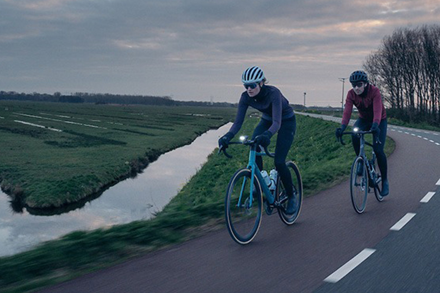 Two cyclists riding road bikes along a winding country road beside a canal during early morning light. 