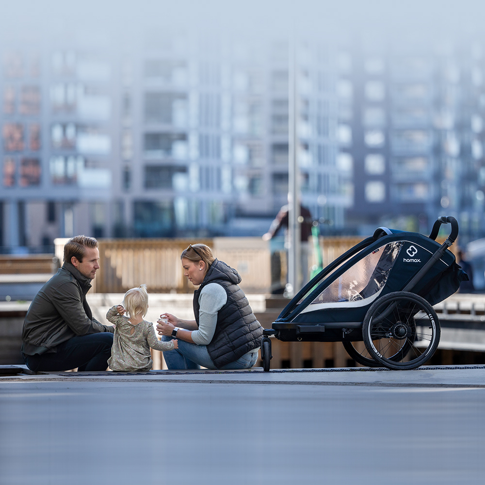 Family enjoying outdoor time with a Hamax bike trailer, seated on a waterfront with urban buildings in the background