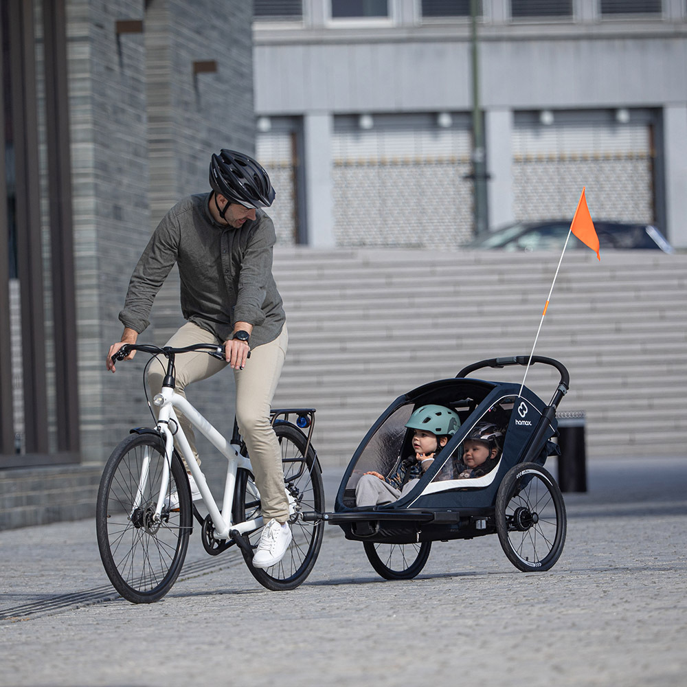 Man riding a bicycle with a Hamax bike trailer attached, carrying two young children with helmets in an urban setting