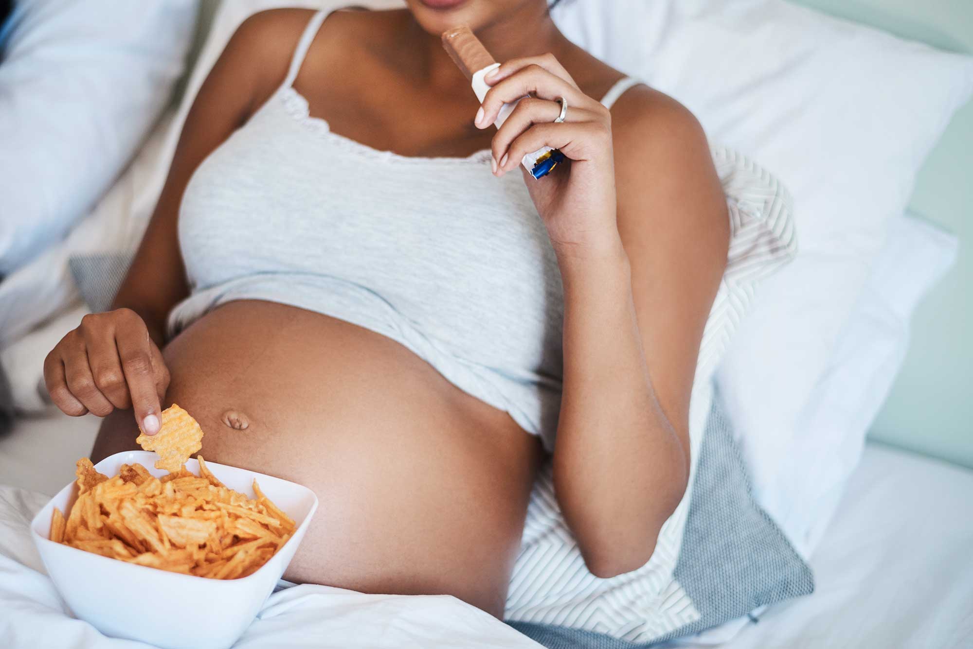Pregnant woman lounging  in bed eating a chocolate bar and potato chips