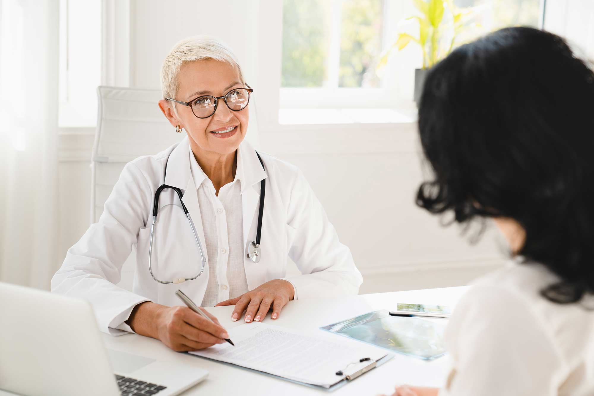 Doctor with a stethoscope having a consultation with a patient, smiling and taking notes at a desk