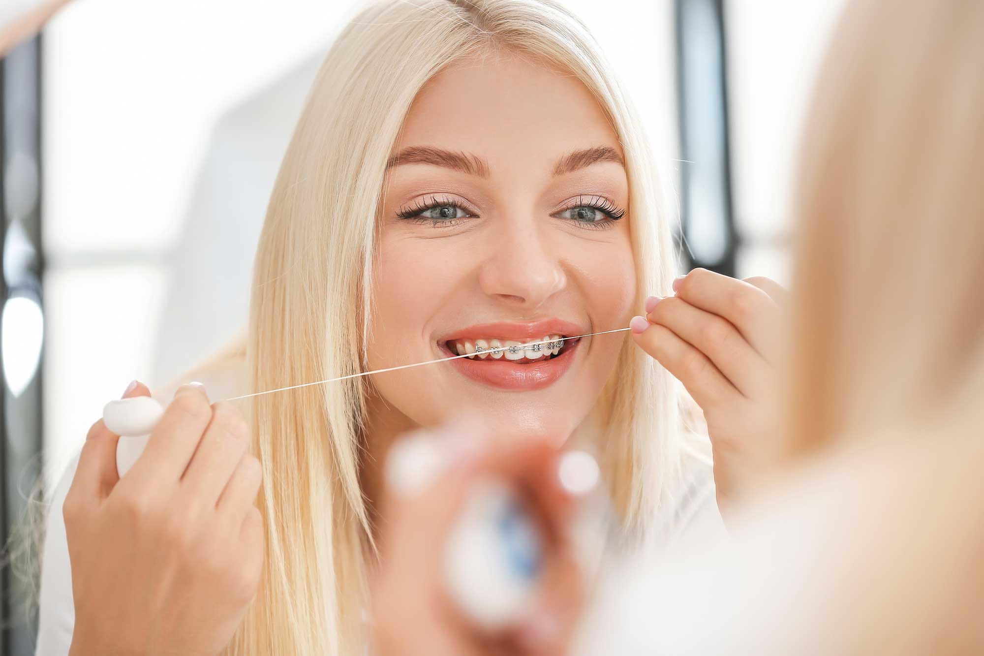Young woman in braces using floss in a mirror Banner Image Young woman in braces using floss in a mirror