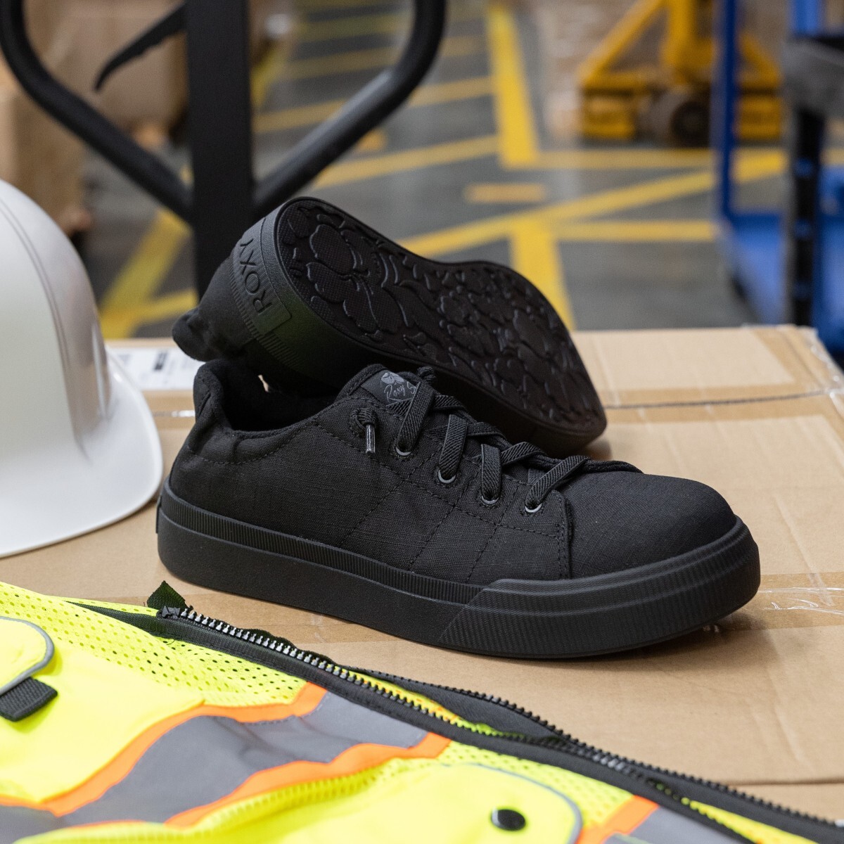 rx90201-rae-work-lifestyle-off-foot-2217-square.jpg Roxy metal free work shoes in style RX90201 sitting on top of a cardboard box next to a hard hat and safety vest with warehouse floor in the background