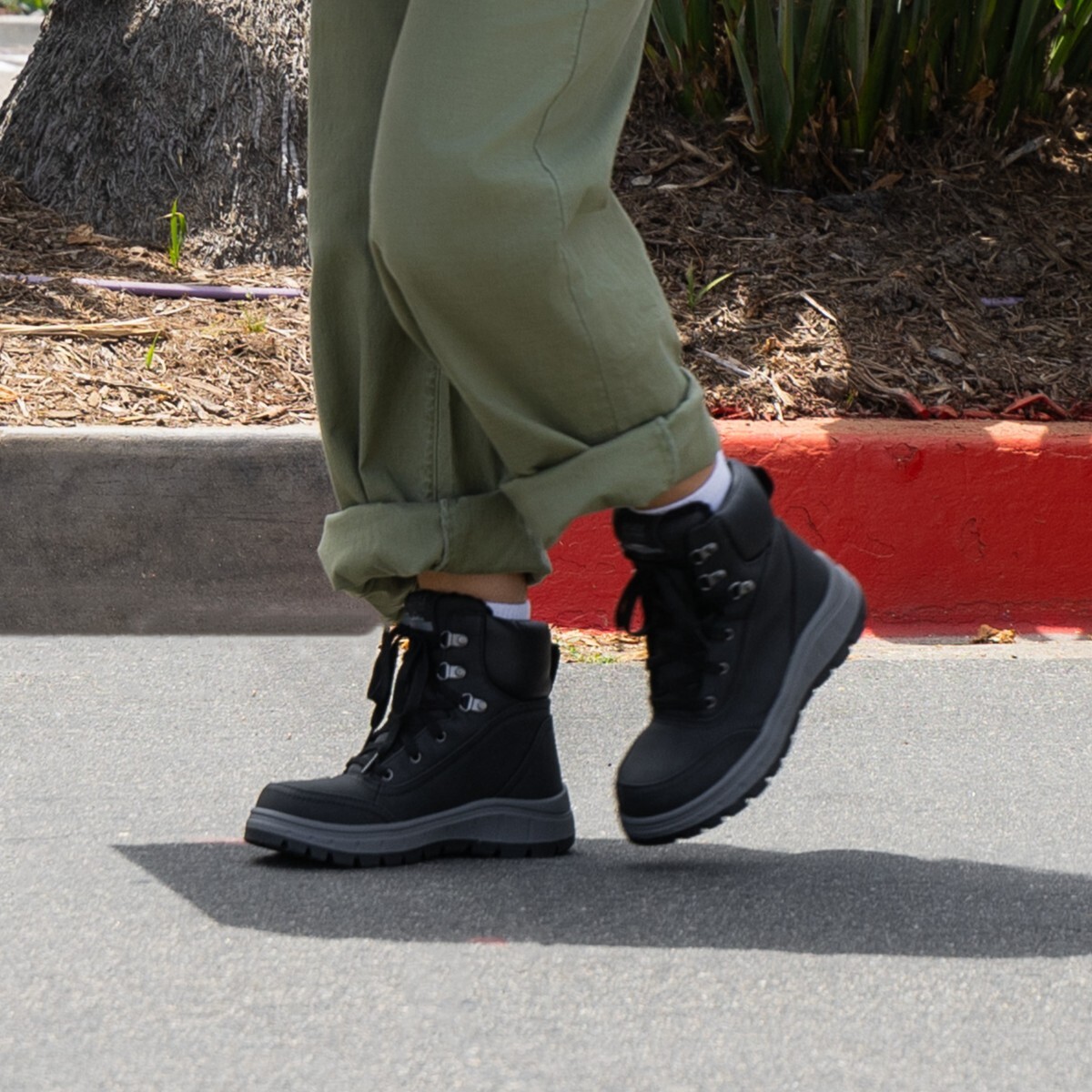  A person wearing rolled-up olive green pants, white socks, and black lace-up hiking-style boots walks on an asphalt surface near a red curb and landscaped area