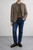 Man wearing Marselo brown herringbone blazer with olive crew‑neck top, dark jeans, and brown loafers against a light textured wall.