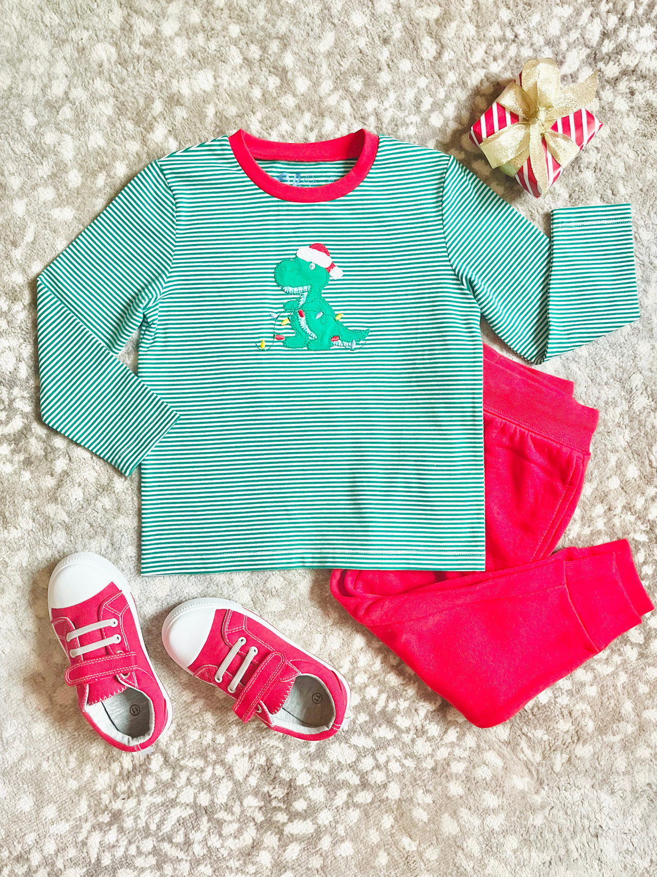 green gingham dinosaur applique on green and white stripe knit shirt with red neck trim. dinosaur is holding embroidered christmas lights. shirt is shown with red pants and red shoes for outfit inspiration and styling.