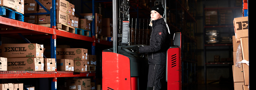 Woman driving a forklift in a cold storage warehouse wears insulated bibs and a work coat.