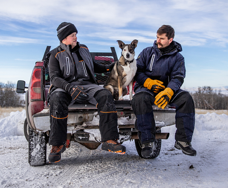 2 Men sitting on the back of a truck in the snow with a dog wearing RefrigiWear to keep warm