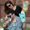 A woman takes a picture with a mobile phone of a pizza-eating guy wearing a Blue Q oven mitt in the "Pizza's Here" style.