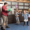 A kitchen scene with a man wearing a Blue Q "Pizza's Here" oven mitt, carrying 3 pizza boxes, and two excited girls.