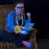 Lady in an armchair, eating, and holding her hot plate with a Blue Q oven mitt with the slogan "Most Likely to Microwave".