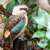 Close-up, high-detail shot of a Kookaburra DecoBird sculpture with vivid blue wing feathers, set against a soft-focus background of green foliage
