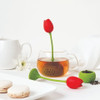 Lifestyle scene showing the red OTOTO Tulip tea infuser steeping in a glass teacup beside macarons and a white teapot.