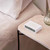 Lexon Flip Classic Alarm Clock in White with 'ON' side up, displayed on a pink marble bedside table in a bedroom setting.