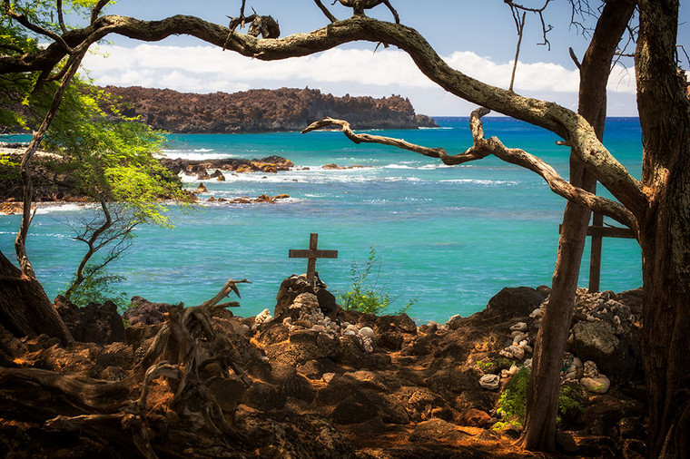 Sunlit shady shoreline with a cross memorial dedicated to surfers at La Perouse Bay in Maui, Hawaii. Turquoise ocean and lava field in background.