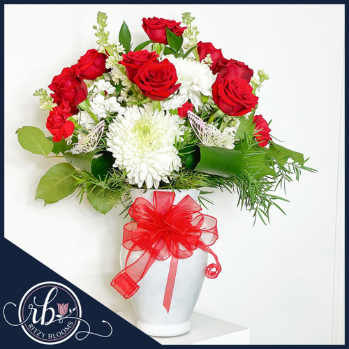 Valentine's Day flower arrangement showcasing vibrant red roses, white chrysanthemums, lush greenery, and butterfly accents in a white vase with a red ribbon.