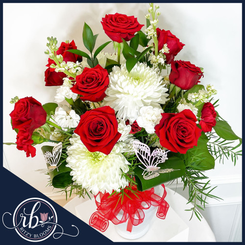Valentine's Day flower arrangement showcasing vibrant red roses, white chrysanthemums, lush greenery, and butterfly accents in a white vase with a red ribbon.