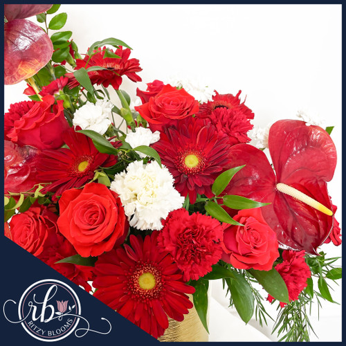 A luxurious floral arrangement of red anthuriums, velvety roses, vibrant gerberas, and white carnations in a gold vase, accented with lush greenery