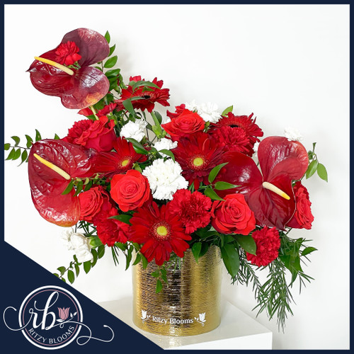 A luxurious floral arrangement of red anthuriums, velvety roses, vibrant gerberas, and white carnations in a gold vase, accented with lush greenery