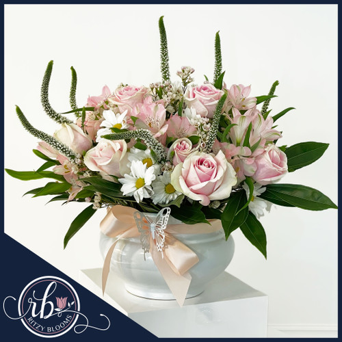 Blush Elegance Baby Floral Arrangement with pink roses, daisies, greenery in a ceramic vase, featuring a satin bow and butterfly accent.