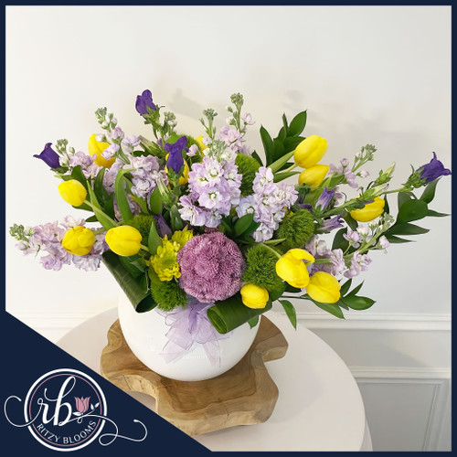 Springtime Symphony Flower Arrangement with lavender stock, yellow tulips, green dianthus, purple campanula, and Pompon Sweet Lavender Button mums in a white vase, designed by Ritzy Blooms. Springtime Symphony Flower Arrangement with lavender stock, yellow tulips, green dianthus, purple campanula, and Pompon Sweet Lavender Button mums in a white vase, designed by Ritzy Blooms.